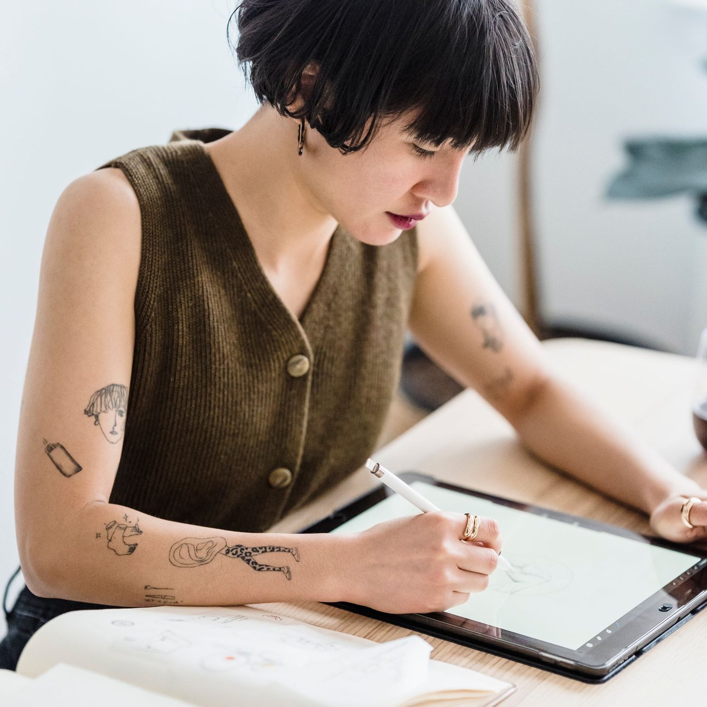 A student with short dark hair writing on a tablet with a stylus at a wooden desk, an open paper notebook beside it
